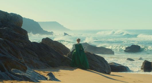 Still from Portrait of a Lady on Fire: a woman in a green dress stands on a rocky beach looking out at waves breaking on the shore.