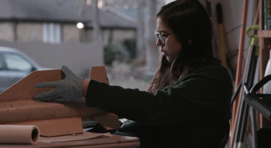 Still from Rocket Girl: a woman in glasses, seen in profile, works at a workbench with a gloved hand on a cardboard tube.