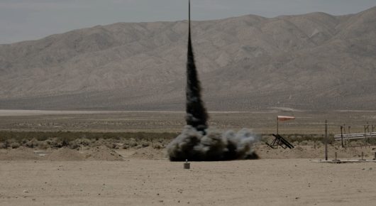 Still from Rocket Girl: a tall dark rocket rises from a puff of smoke on a flat desert, with mountains behind.
