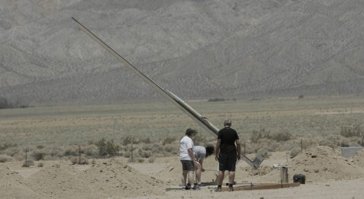 Still from Rocket Girl: two people stand beside a long launch rail angled into the sky in a desert.
