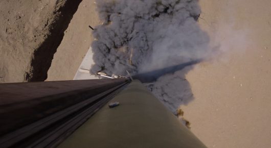Still from Rocket Girl: a view along a long metal launch tube from above, with grey smoke billowing beside it.