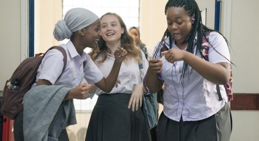 Still from Rocks: three schoolgirls in uniform walk along a corridor laughing, one whispering to another.