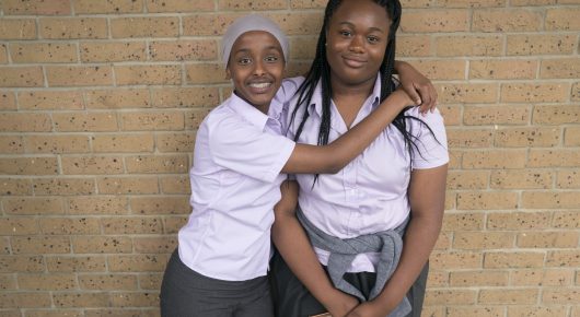 Still from Rocks: two schoolgirls in white shirts hug and smile in front of a brick wall.