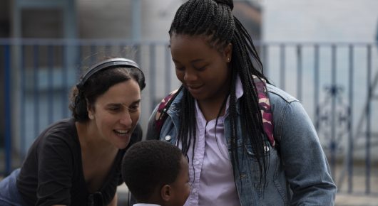 Still from Rocks: a girl with long braids in a denim jacket looks down at a child, with another girl beside her, in front of railings.