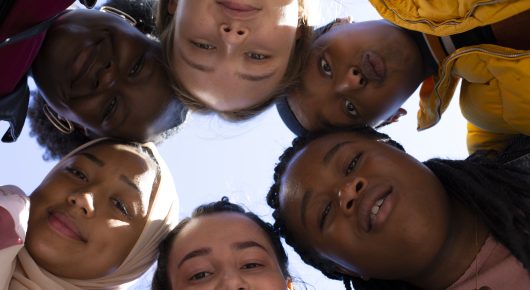 Still from Rocks: a view from below of five girls leaning in with their faces close together and smiling, some in headscarves.