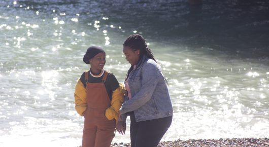 Still from Rocks: two girls stand in shallow, sparkling water at the edge of the sea.