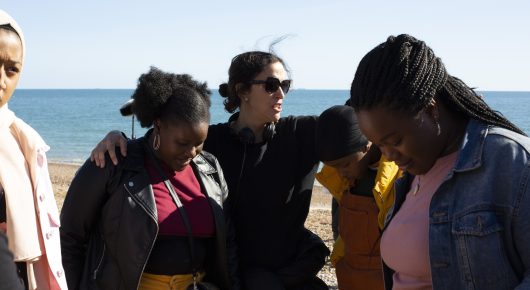 Still from Rocks: a group of girls stand together on a beach with their arms around each other, the sea behind them.