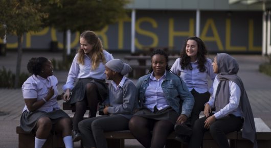 Still from Rocks: a group of schoolgirls sit on a bench outside a building, talking and laughing.