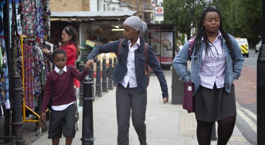 Still from Rocks: two girls walk along a busy street past a market stall of fabrics, with a child in a school uniform beside them.