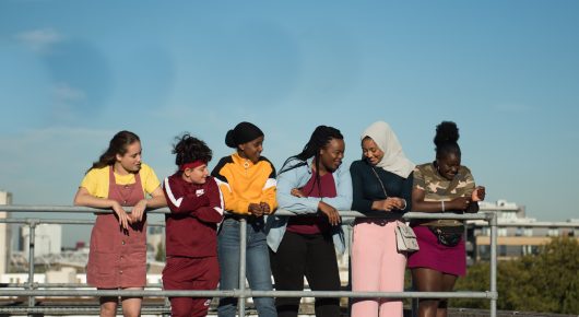 Still from Rocks: seven girls stand in a row along a rooftop railing, looking out, with a city behind them.