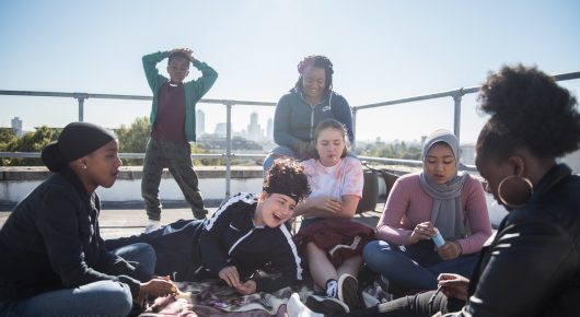 Still from Rocks: a group of girls sit and lie on a rooftop, with one standing on a ledge behind them.