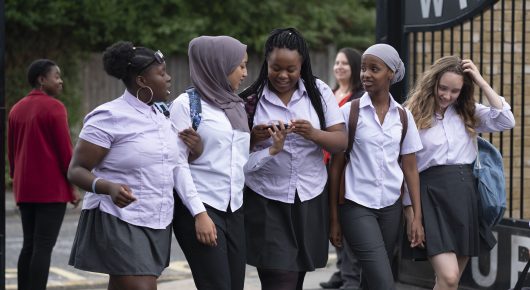 Still from Rocks: a group of teenage girls in school uniform, some in headscarves, walk together through school gates.