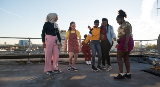 Still from Rocks: a group of girls stand and talk on a rooftop car park with a city skyline behind them.