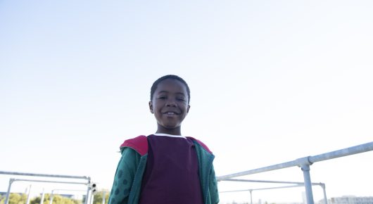 Still from Rocks: a girl in a teal and purple jacket, seen from below against a pale blue sky.