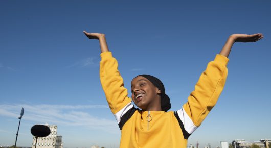 Still from Rocks: a girl in a yellow jacket raises both arms against a blue sky.