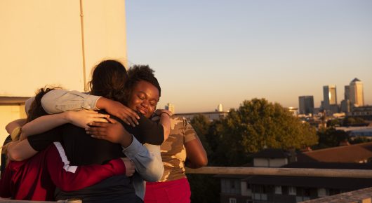 Still from Rocks: girls hug on a rooftop at sunset, with city buildings behind them.