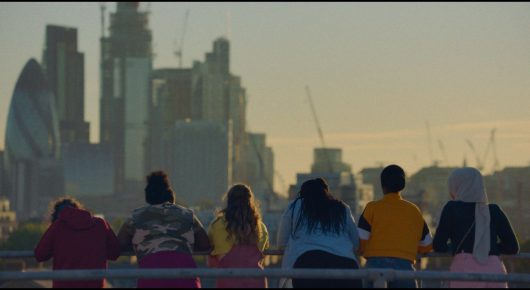 Still from Rocks: a group of young people sit and stand at a railing looking out over the London skyline at dusk, seen from behind.