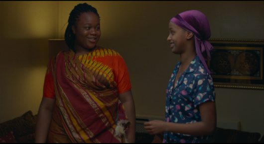 Still from Rocks: a woman in an orange sari-style outfit and a girl in a purple headscarf and floral top stand talking in a dimly lit room.