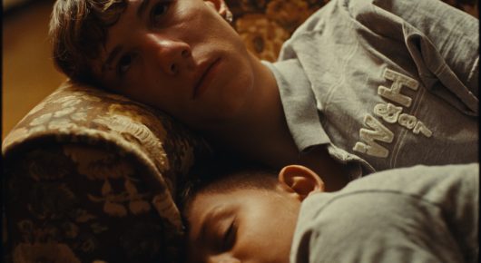 Still from Rage: a young man in a grey sweatshirt lies on a patterned sofa with a smaller person curled against him.