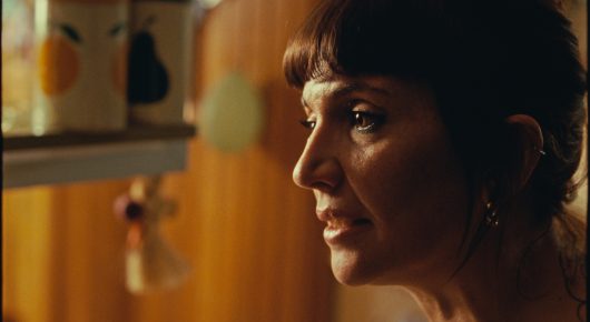 Still from Rage: a woman with dark hair and a fringe looks to one side in a warmly lit kitchen.