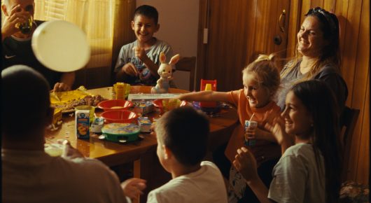 Still from Rage: a family sits around a crowded dining table with food and a balloon, in a warmly lit room.