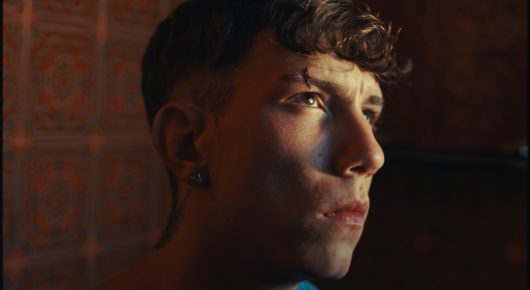 Still from Rage: a close-up side view of a young man with short dark hair, lit from one side, looking off to the side.