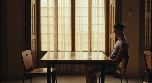 Still from Rage: a young man sits alone at a table in a plain room with tall windows letting in light.