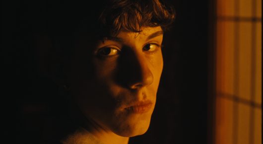 Still from Rage: a close-up of a young person with tousled dark hair looking at the camera from the shadows, lit with orange light.