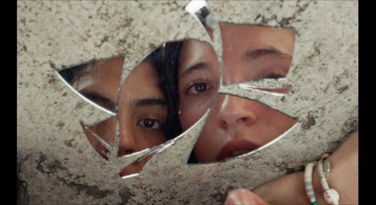 Still from Sad Girlz: a cracked mirror shows fragments of a young woman's face against a pale stone wall.
