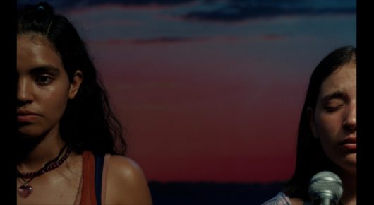 Still from Sad Girlz: two young women stand side by side at dusk, one looking at the camera and one with her eyes closed.