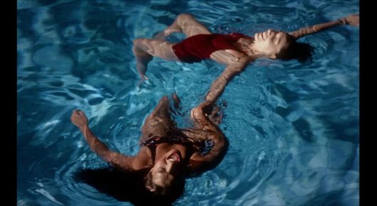 Still from Sad Girlz: people float in blue water, seen from above, one wearing red.
