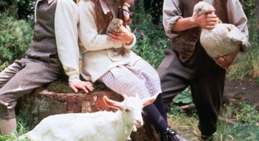 Still from The Secret Garden: two children sit on a tree stump with a white goat in front of them, one holding a white bird.