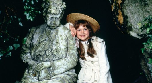Still from The Secret Garden: a girl in a straw hat and white coat stands beside a moss-covered stone statue in a dark garden.