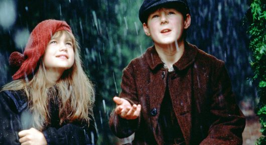 Still from The Secret Garden: a girl in a red hood and a boy in a dark flat cap and red-brown jacket stand in the rain, the boy holding out his palm.
