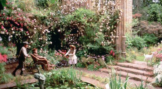 Still from The Secret Garden: a sunlit garden full of pink roses and flowering shrubs, with stone steps and a few small figures walking among the flowers.