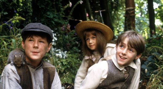 Still from The Secret Garden: a boy in a flat cap with a squirrel on his shoulder, a girl in a straw hat and a boy in a waistcoat sit close together in a leafy garden.