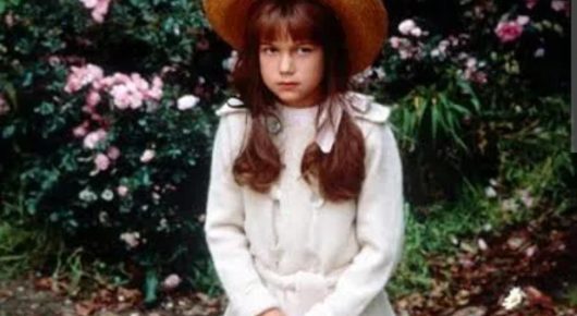 Still from The Secret Garden: a girl with long brown hair in a straw hat and white coat stands in a garden with pink flowering bushes behind her.