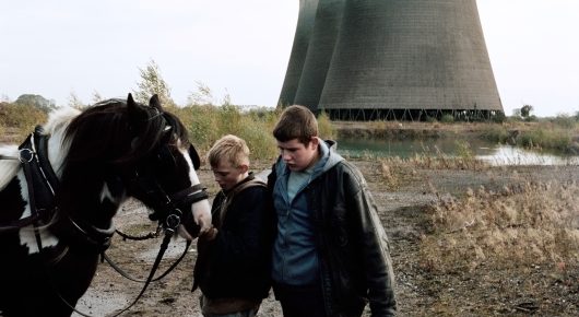 Still from The Selfish Giant: two boys stand beside a black and white horse on waste ground, with a power-station cooling tower behind them.