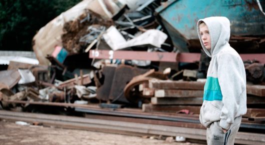 Still from The Selfish Giant: a boy in a white and teal hooded top stands beside a pile of scrap metal in a scrapyard.