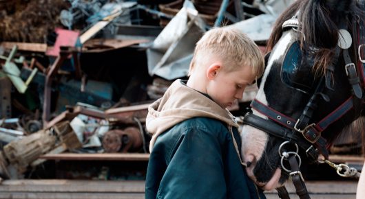 Still from The Selfish Giant: a blond boy in a hoodie stands with his head bowed beside a horse in a scrapyard full of metal.