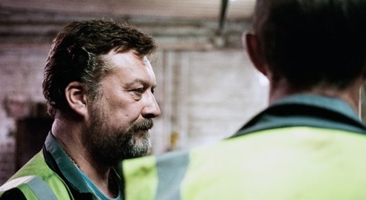 Still from The Selfish Giant: a bearded man in a high-visibility jacket looks to the side, with the back of a boy's head in the foreground.