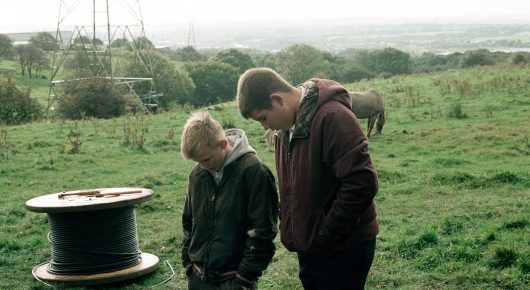 Still from The Selfish Giant: two boys in dark jackets stand on grass beside a large cable reel, with pylons and a horse in the distance.