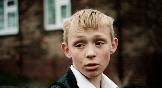 Still from The Selfish Giant: a close-up of a blond boy in a school shirt and tie looking up with a worried expression, in front of a brick building.