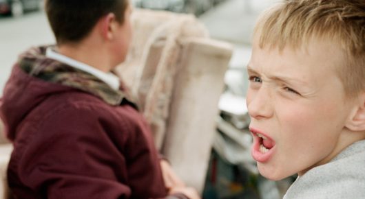 Still from The Selfish Giant: a blond boy with his mouth open speaks beside a boy in a maroon hoodie seen from behind, with an armchair behind them.
