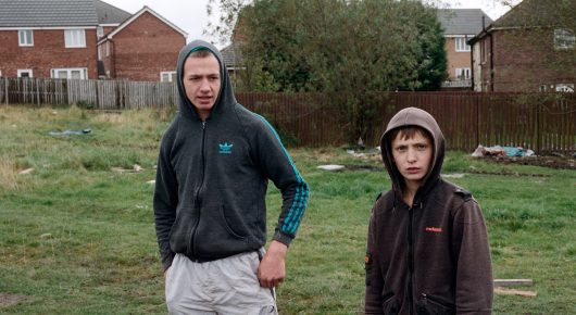 Still from The Selfish Giant: two boys in hooded jackets stand on a patch of grass in front of terraced houses and a fence.