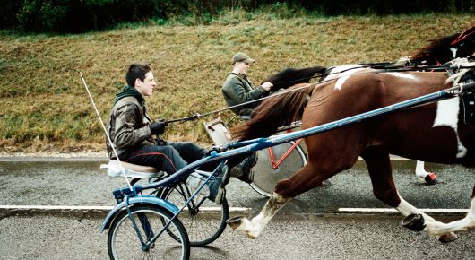 Still from The Selfish Giant: a person in a dark jacket drives a brown and white horse pulling a light two-wheeled cart along a road, with another cart behind.