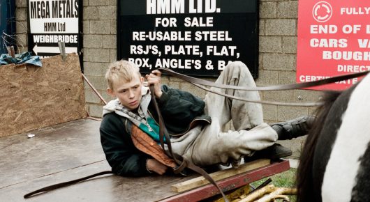 Still from The Selfish Giant: a boy lies on his back on the ground beside a horse, in front of a scrapyard sign.