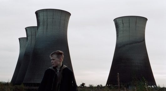 Still from The Selfish Giant: a boy stands between two huge power-station cooling towers under a grey sky.