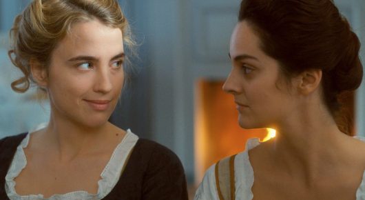 Still from Portrait of a Lady on Fire: two women in 18th-century dress stand close together in a room, one looking at the other with a faint smile.