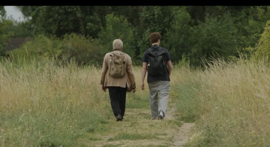 Still from Cambridge Connections: an older woman and a younger person with a rucksack walk away along a path through tall grass.